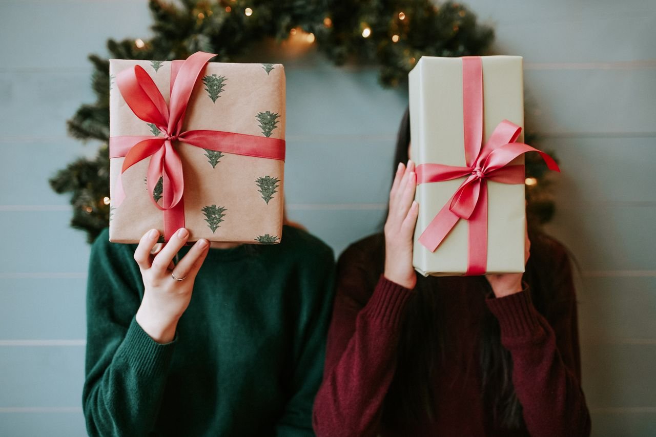 Two people holding Christmas gifts in front of a festive wreath indoors.